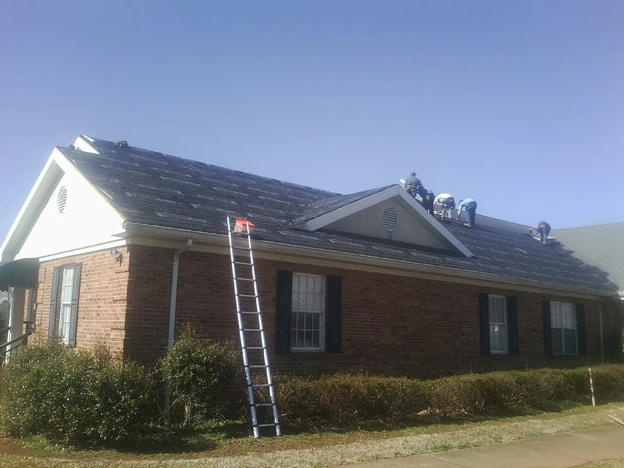 Crew tearing shingles off a brick ranch roof with a ladder set — Emergency Roof Repair work by our crew in Eagle Creek