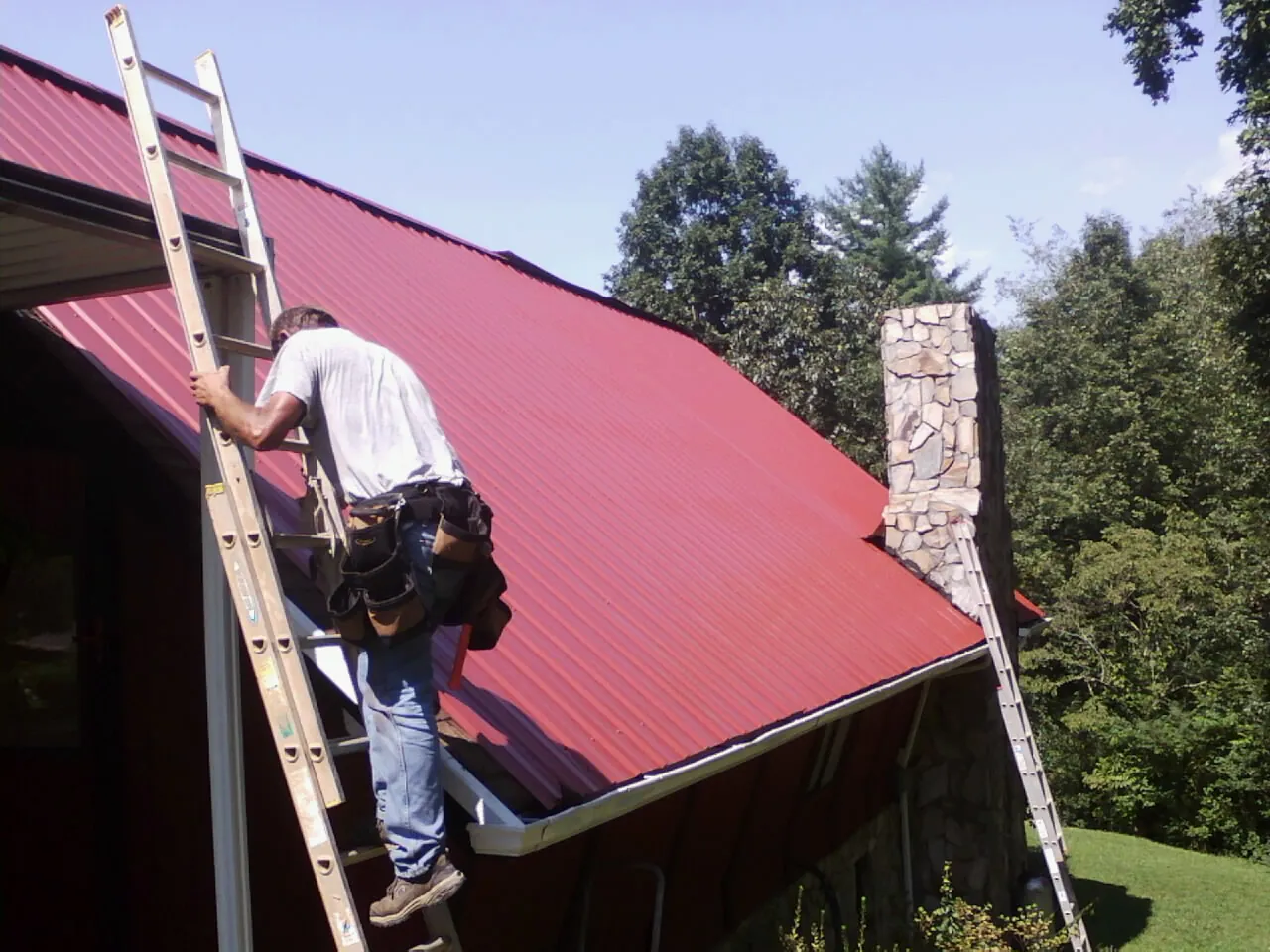 Roofer on a ladder beside a red metal roof and stone chimney — Soffit & Fascia Repair by Roofing Brothers in Eagle Creek